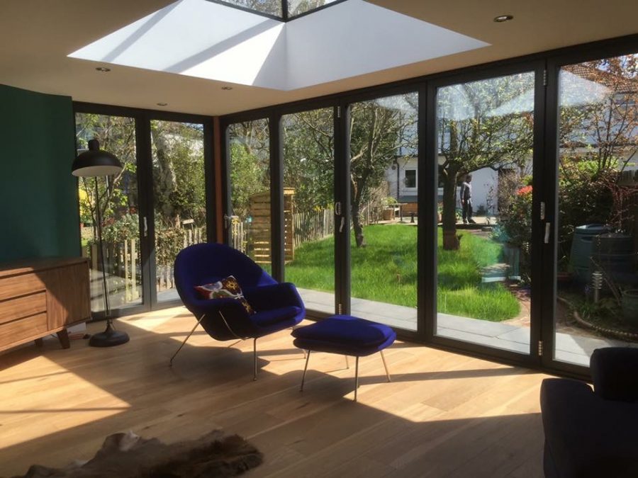 extension with multi-panel glass doors leading to garden with blue cushioned chair under pyramid roof lantern.