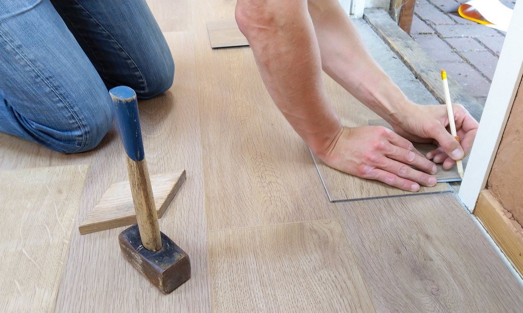 Man on his knees next to a hammer measuring the floor and wall with a pencil.